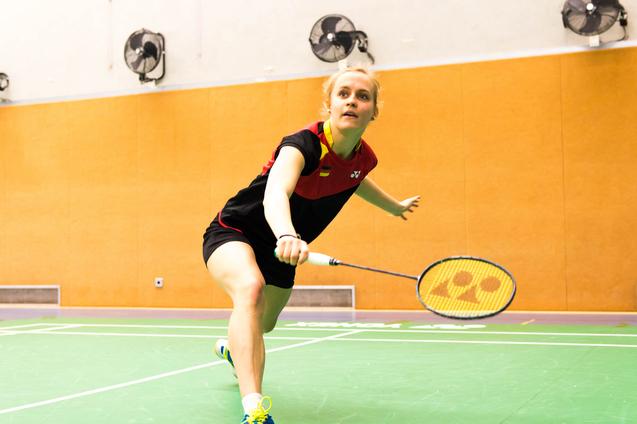 Spielerin schlägt einen Badmintonball auf einer grünen Spielfläche in einer Sporthalle mit orangefarbenen Wänden.