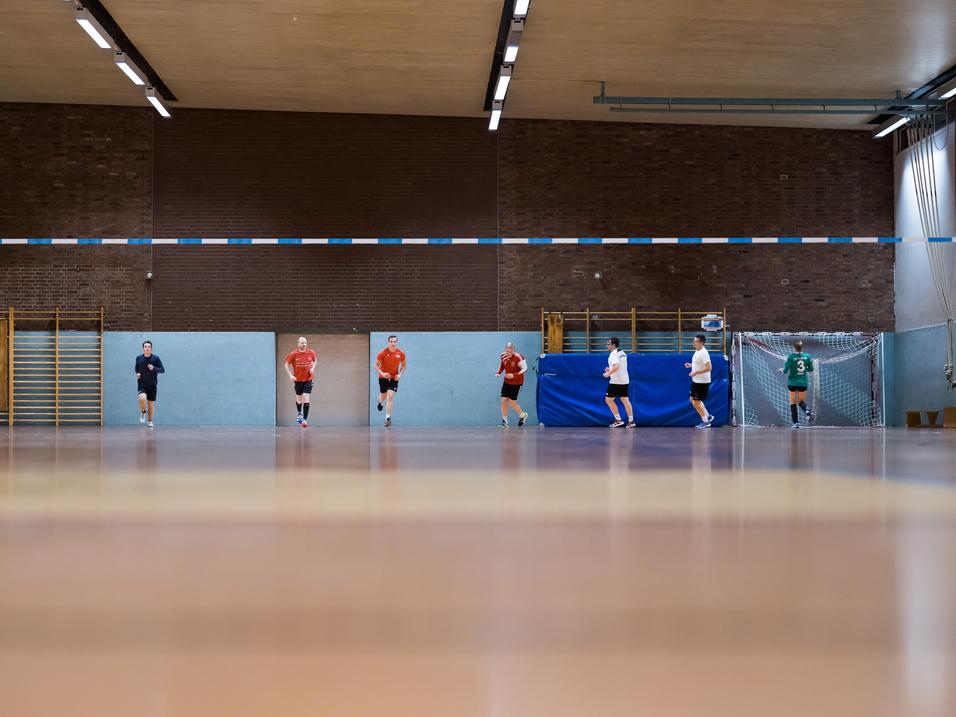 Fünf Spieler in Sportkleidung trainieren in einer Sporthalle, mit einem Tor und blauen Wänden im Hintergrund.