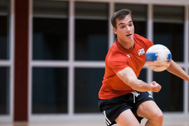 Junger Sportler in rotem Shirt fängt einen Handball in einer Sporthalle mit Holzboden.