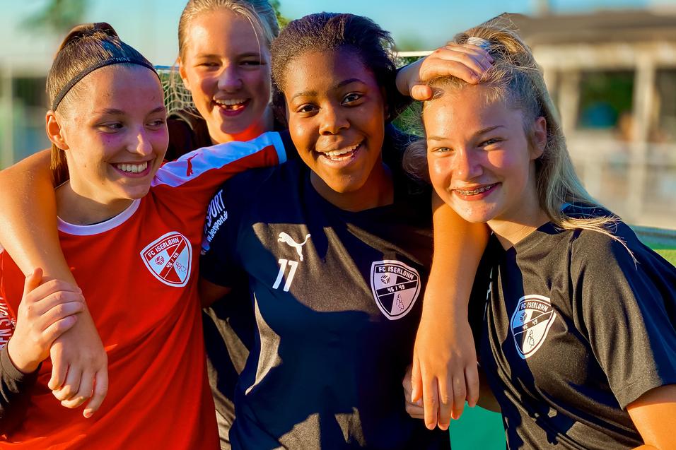 Vier junge Fußballspielerinnen in Teamtrikots posieren lachend gemeinsam auf dem Spielfeld.