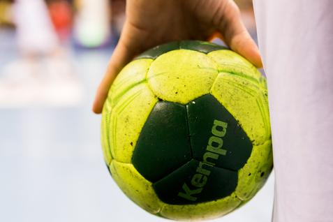 Nahaufnahme einer Hand, die einen grünen Handball mit gelben Elementen hält, im Hintergrund Spieler auf dem Spielfeld.