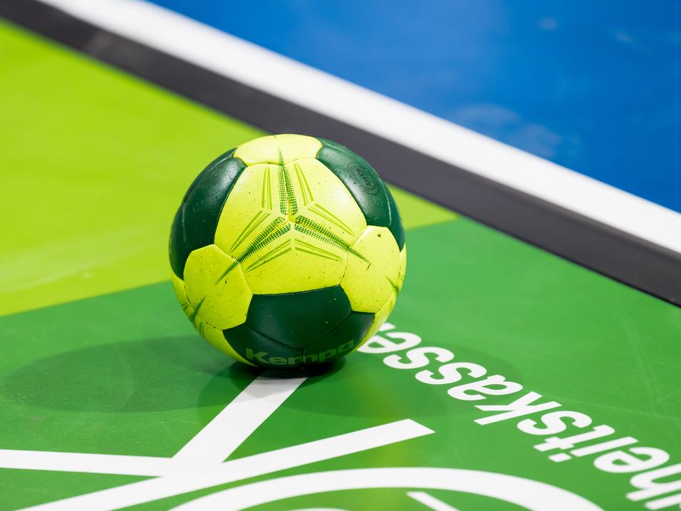 Grüner und gelber Handball liegt auf einer grünen Spielfläche mit weißem Schriftzug.