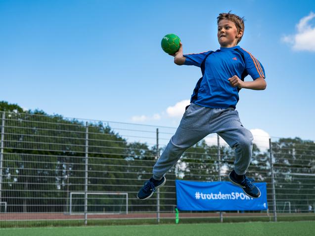 Junge springt auf einem Sportplatz und wirft einen grünen Handball, umgeben von einem Zaun und Bäumen im Hintergrund.