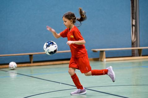 Mädchen in rotem Trikot dribbelt einen Fußball in einer Halle mit grünem Boden und blauen Wänden.