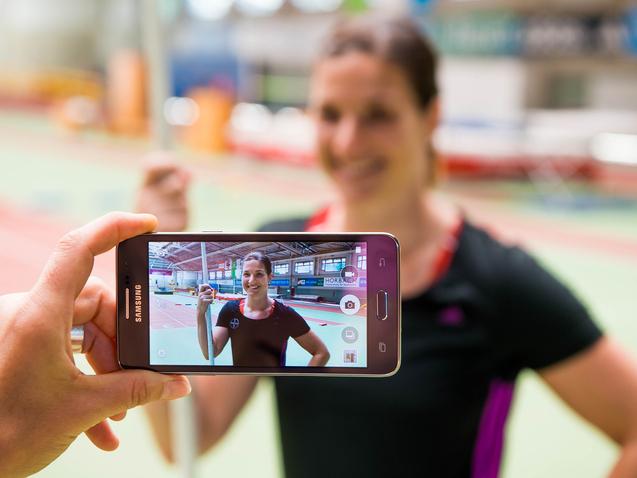 Hand hält ein Smartphone, das ein Porträt einer lächelnden Athletin mit Stab in einer Sporthalle zeigt.