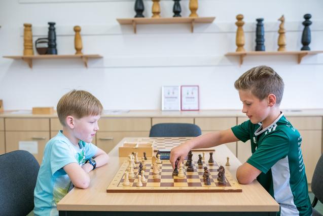 Zwei Kinder spielen Schach an einem Holztisch, mit Figuren auf dem Schachbrett und einer digitalen Uhr.