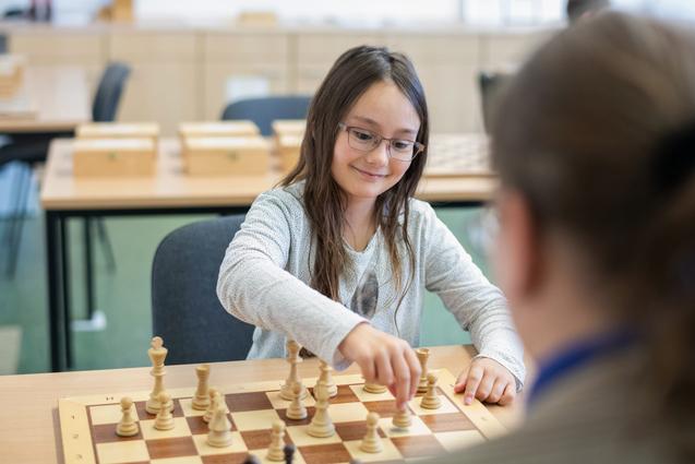 Mädchen mit Brille zieht eine Schachfigur auf einem Schachbrett in einem Klassenraum. Holzschachfiguren im Vordergrund.