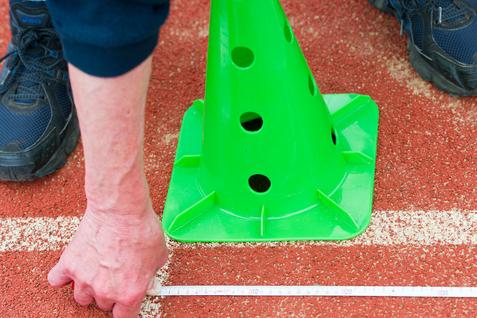 Hand misst mit einem Zollstock die Distanz zu einem grünen Pylonen auf einer roten Sportfläche.