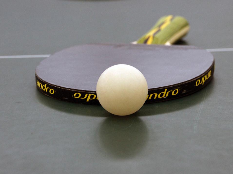 Nahaufnahme eines Tischtennisschlägers mit weißem Ball auf einer grünen Tischtennisplatte.