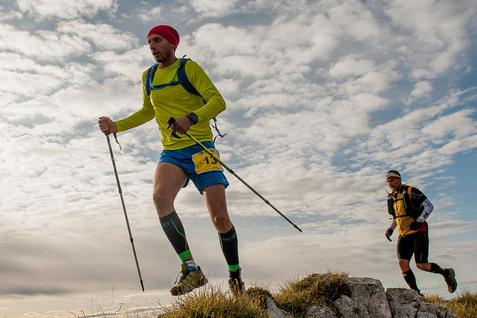 Zwei Männer in Sportkleidung wandern mit Stöcken über einen felsigen Bergrücken unter einem bewölkten Himmel.