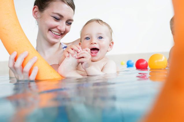 Eine fröhliche Mutter hält ein kleines Kind im Wasser, während sie einen Schwimmring und bunte Bälle um sich haben.
