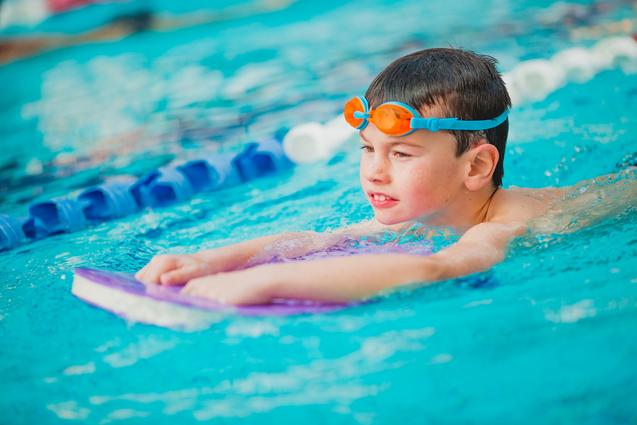 Junge mit orangenen Schwimmbrille übt auf einem Schwimmbrett im Wasser eines Schwimmbeckens.