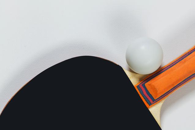 Nahaufnahme eines schwarzen Tischtennisschlägers mit orangefarbenem Griff und einem weißen Ball auf einem weißen Hintergrund.