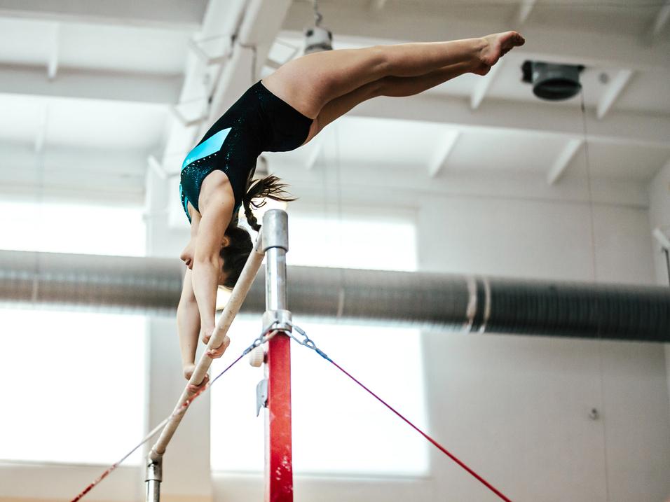 Turnerin in einem schwarzen Trikot führt einen Schwung an den Stufenbarren in einer hellen Turnhalle aus.